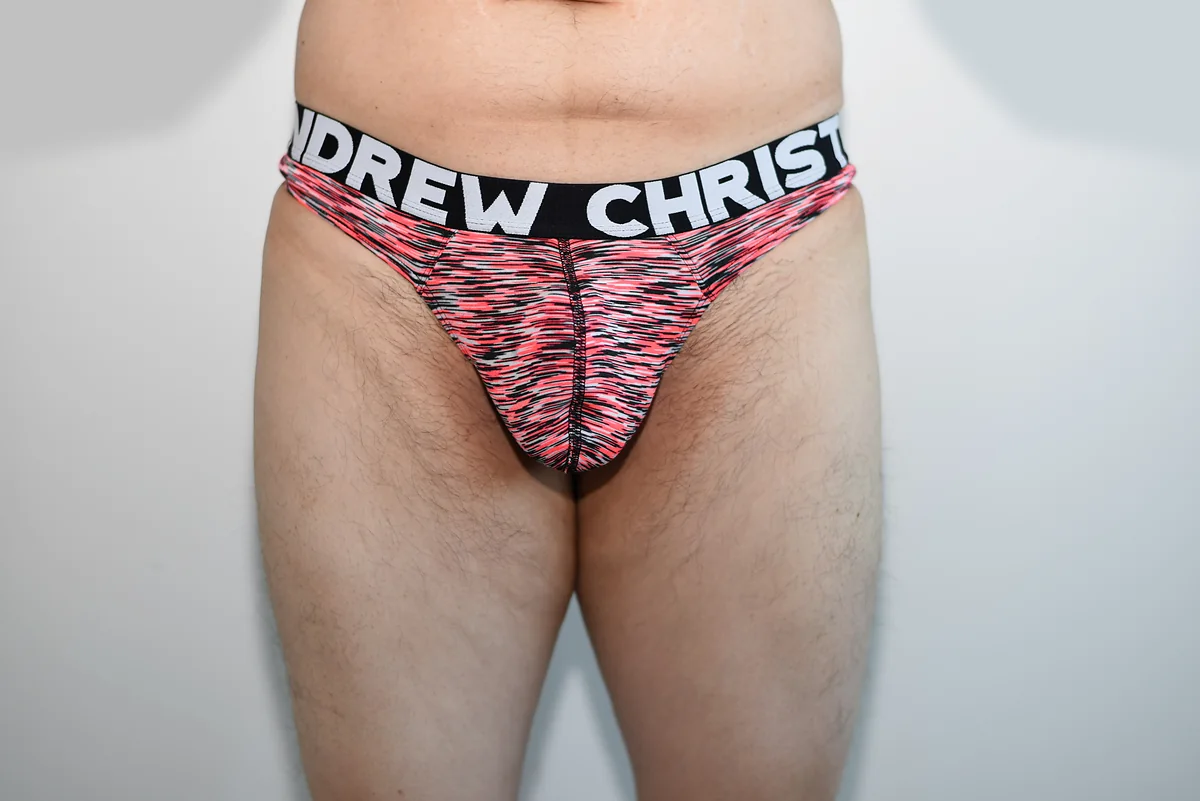 Full front view of the red, black, and white Andrew Christian Happy Gym Thong in size medium, with both upper thighs visible against a neutral wall.