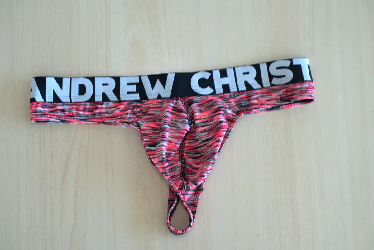 The thong laid flat from the front on a light wood surface, showing the branded waistband, contoured pouch, seams, and lower opening.