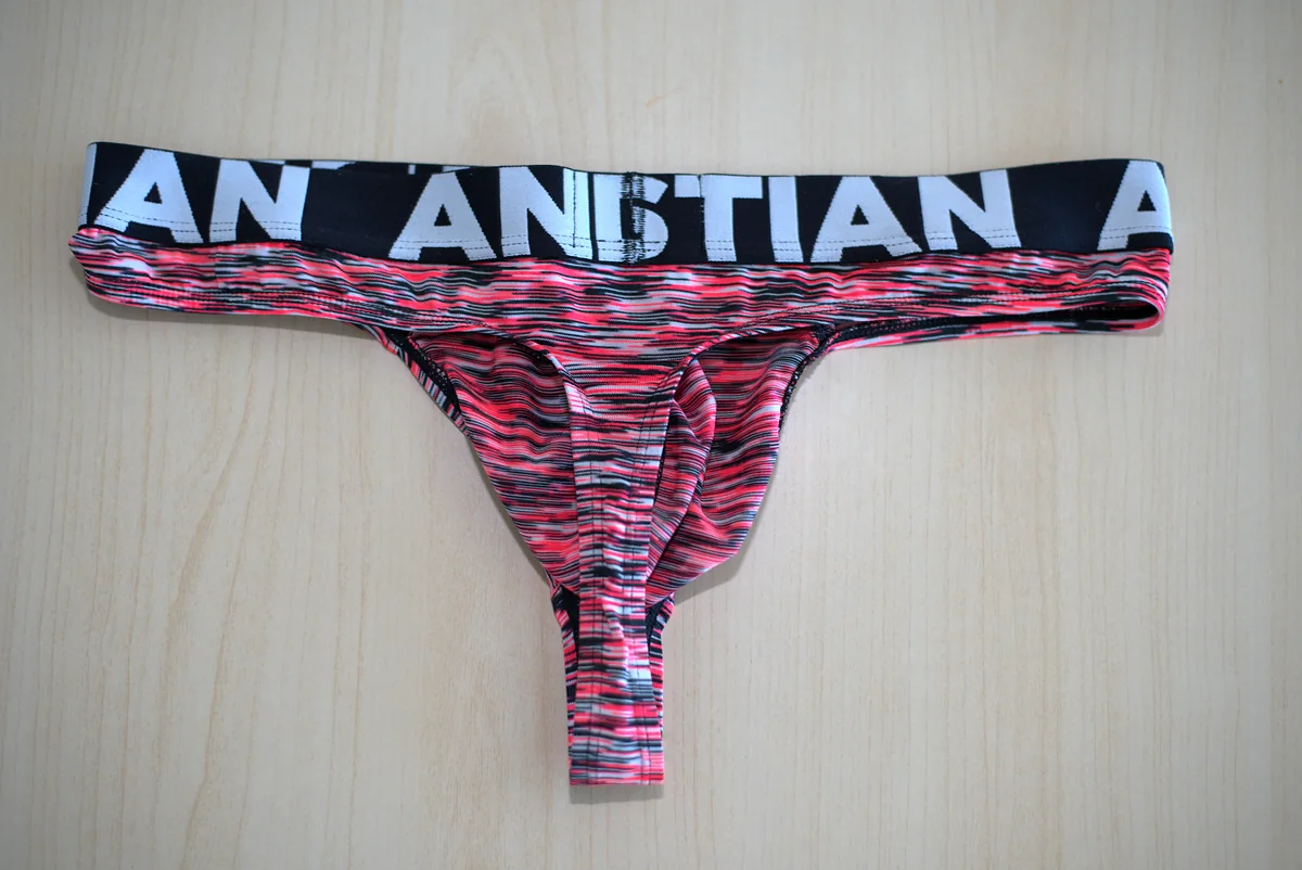 The thong laid flat from the rear on a light wood surface, showing the branded waistband, rear panel, and full narrow tail.