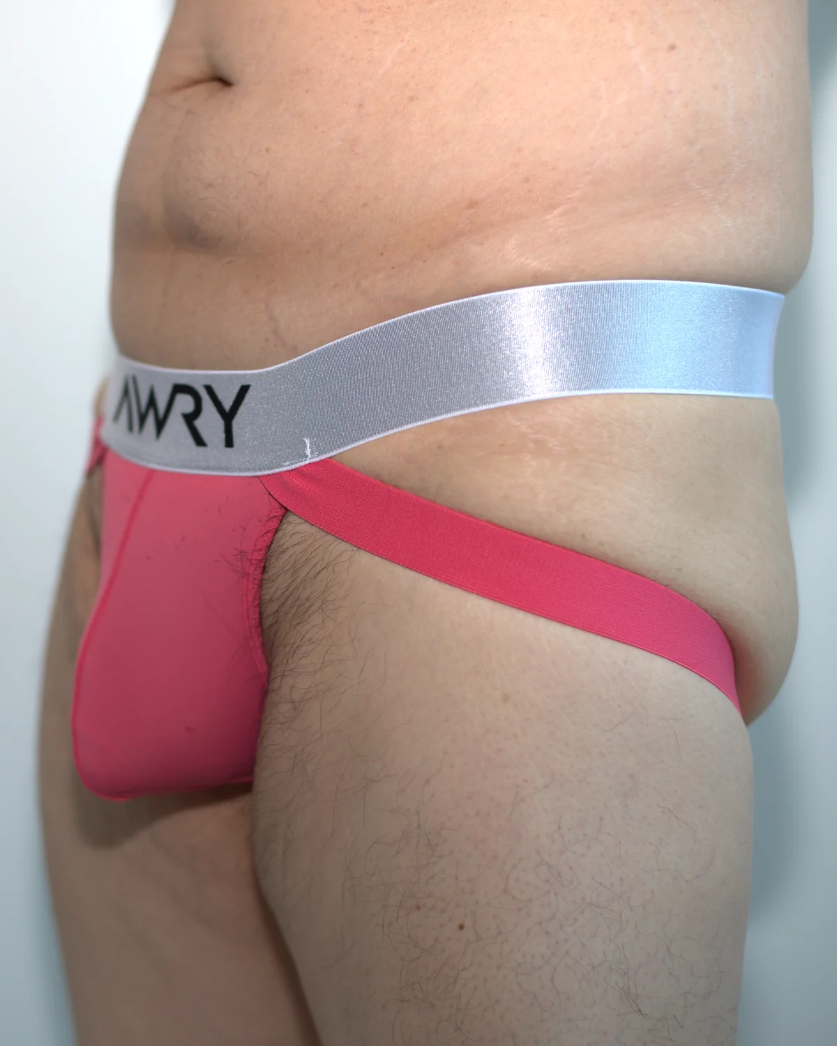 Front three-quarter view of the pink AWRY APEX Jock Thong in Medium, showing the shallow pouch, silver waistband, lower strap placement, and upper thigh.