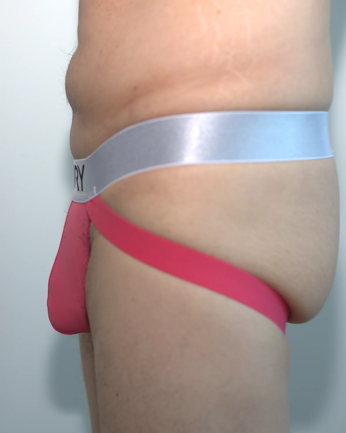 Side view of the pink AWRY APEX Jock Thong in Medium, showing the low pouch profile, silver waistband, lower strap, and open rear.