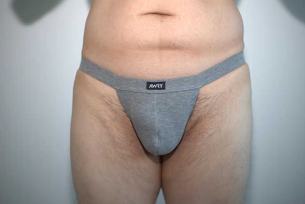 Wide front view of the heather gray AWRY RIBBED Thong in Medium, showing the ribbed pouch, higher-rise waistband, upper thighs, and surrounding neutral wall.