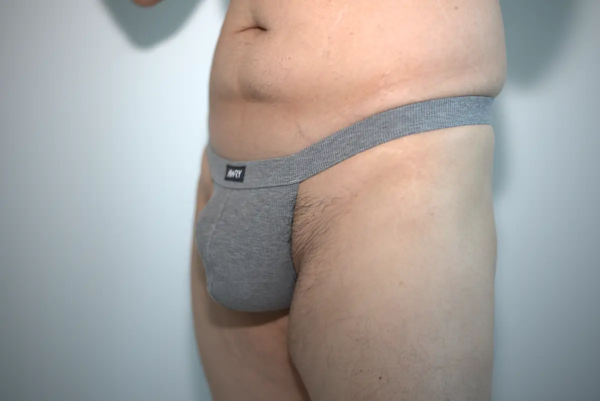 Wide front three-quarter view of the heather gray AWRY RIBBED Thong, showing pouch projection, waistband rise, leg opening, and surrounding neutral wall.