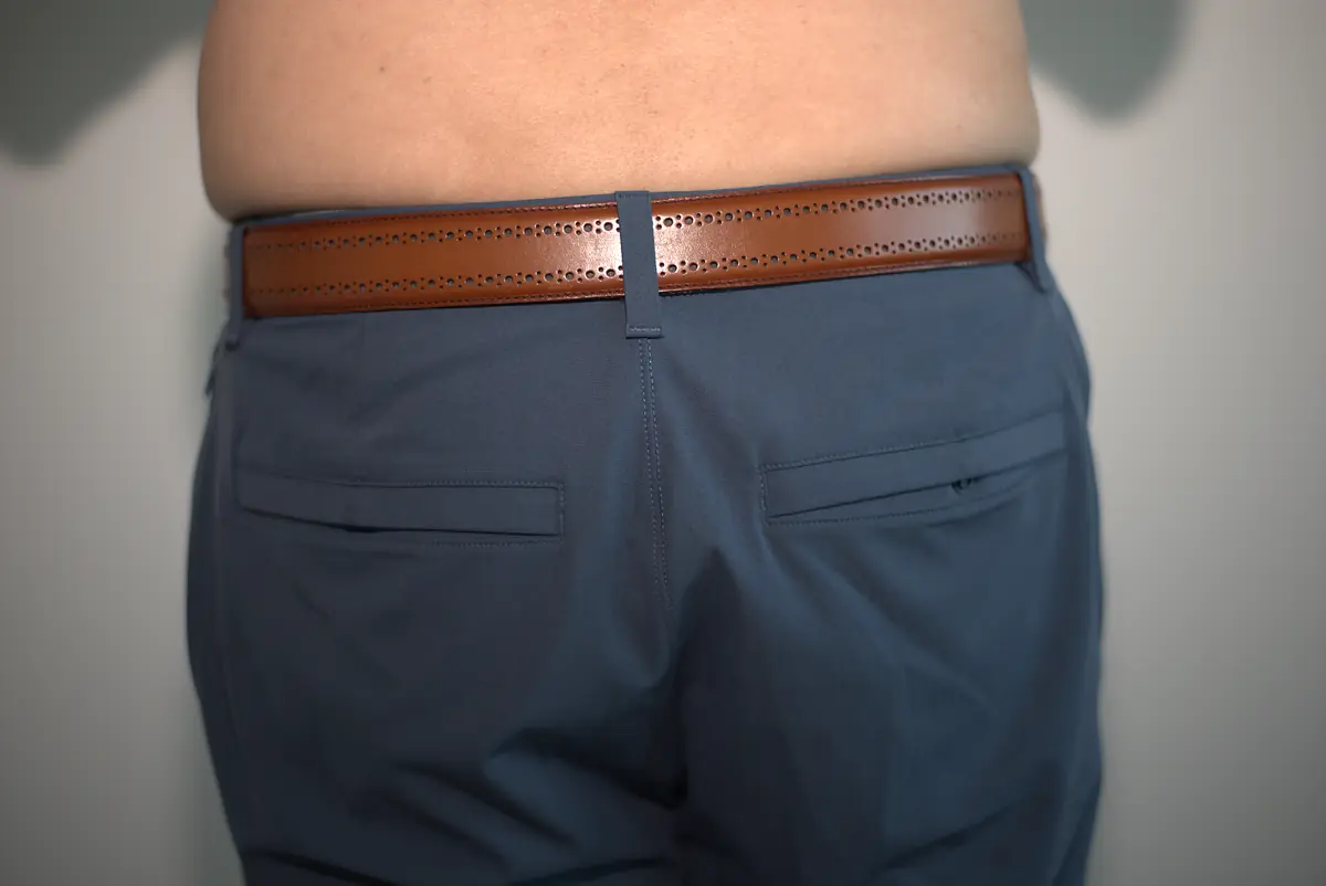 Wide rear view of blue-gray dress pants worn over the AWRY RIBBED Thong, showing a smooth outline beneath the trousers and surrounding neutral wall.