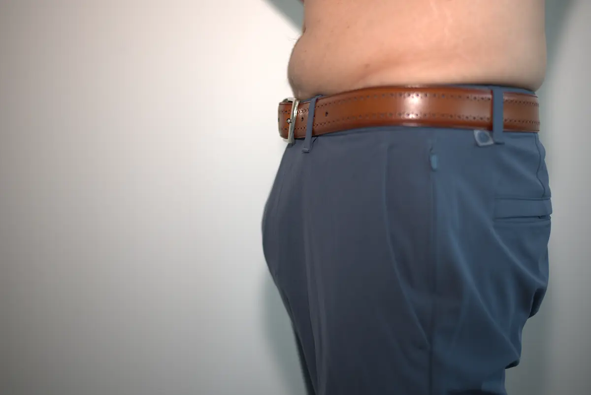 Wide side view of blue-gray dress pants worn over the AWRY RIBBED Thong with a tucked-in shirt, belt, and surrounding neutral wall.