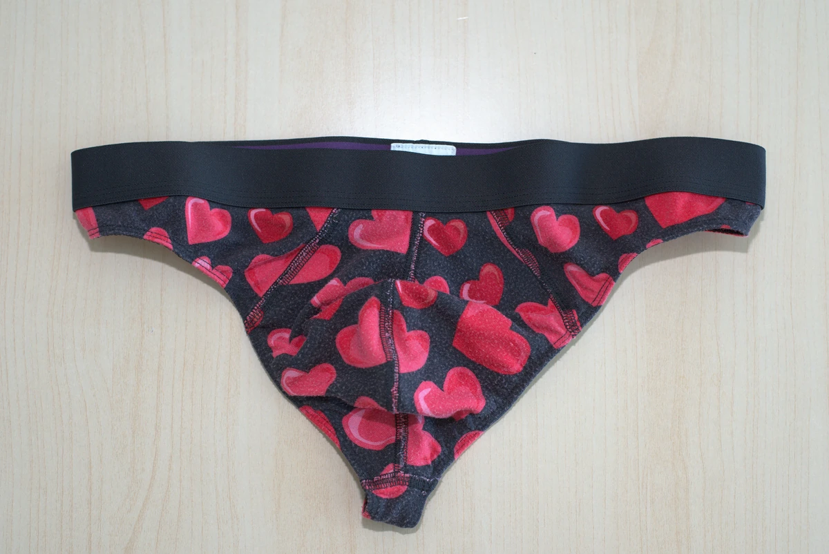Front flat lay of the hearts-print MeUndies Pouch Front Thong on a light wood surface, with the full garment visible.
