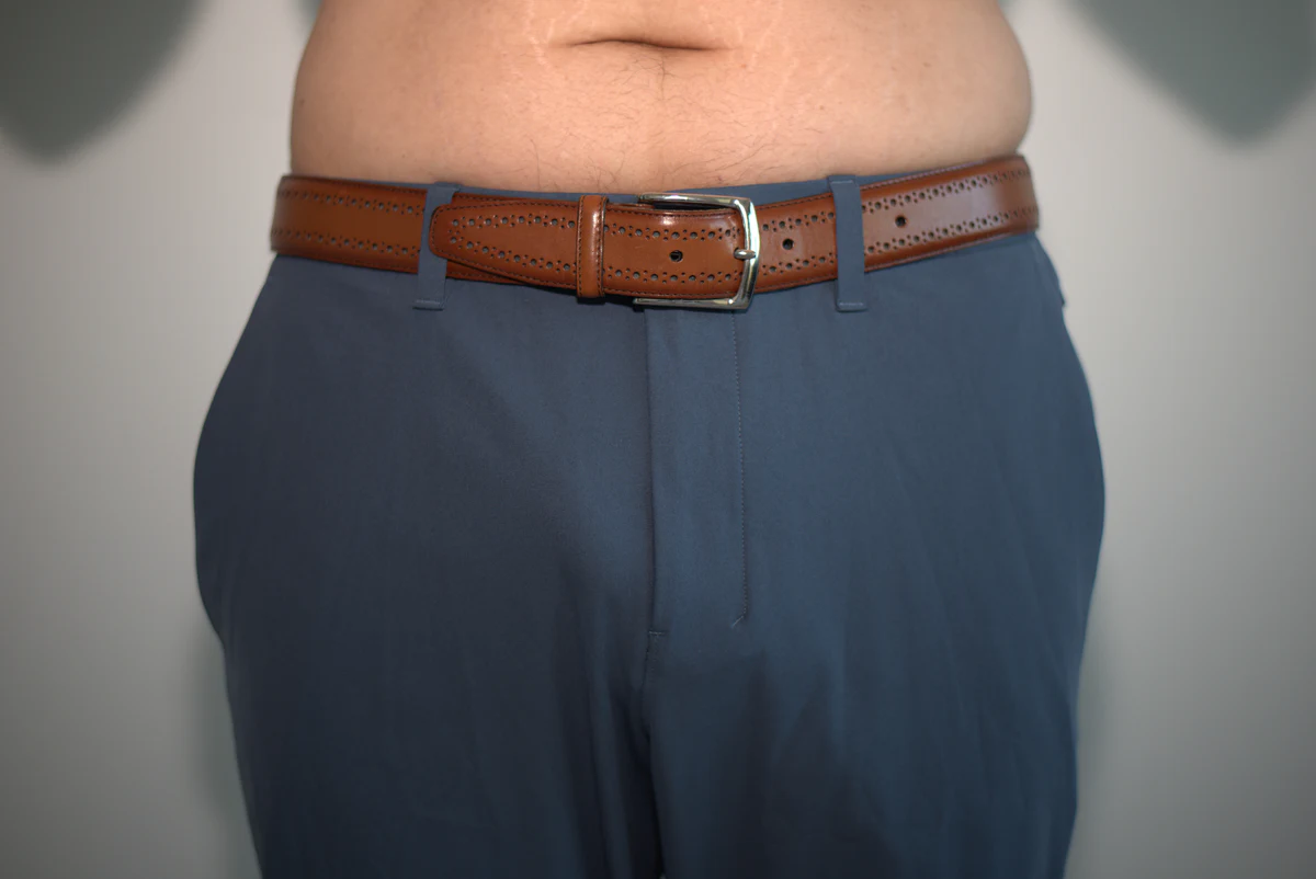 Wider front view of fitted dress pants worn over the Primeman Thong, with both outer edges of the pants and surrounding wall visible.