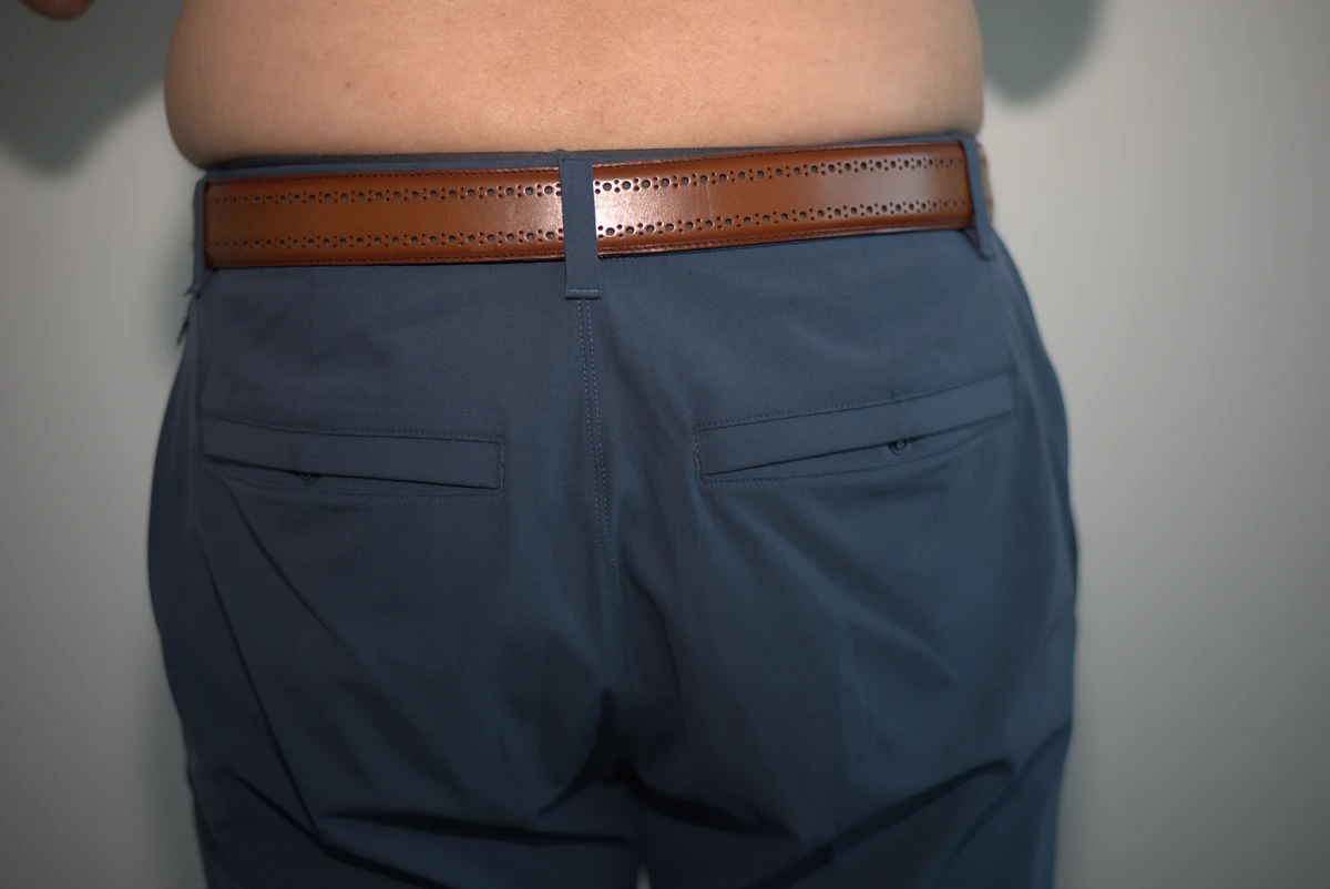 Wider rear view of fitted dress pants showing a smooth finish over the Primeman Thong, with both outer edges of the pants and surrounding wall visible.